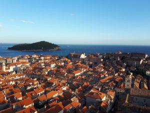 Man sieht Dubrovnik von oben, mit seinen vielen dicht an dicht gebauten Häuser mit ihren orangen Dächern und die davor im Meer liegende Insel.