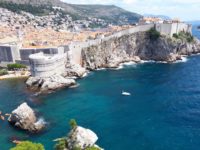 Das Bild zeigt Dubrovnik mit seiner felsigen Küste und der hohen Stadtmauer. Vor Dubrovnik im Mehr sieht man ein paar Kajakfahrer und ein kleines weißes Boot.