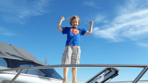 Der Sohn der Familie steht barfuß auf dem Bug der 20 -Yachtline- Er trägt ein blaues Superman T-Shirt und eine karierte Hose. Er stärkt die beiden geballten Fäuste in die Luft und lächelt in die Kamera.