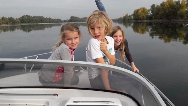 Die 3 kleinsten Kinder der Familie sitzen vorne auf dem Bug des Bootes, das Foto wurde vom Fahrerstand aus gemacht. Alle 3 lächeln in die Kamera.