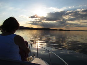 Am linken Rand des Bild sitzt Frau S. auf dem Bug des Boot, die Sonne ist am untergehen und es dämmert schon leicht. Die Sonne bricht durch die Wolken und der Himmel leuchtet orange.