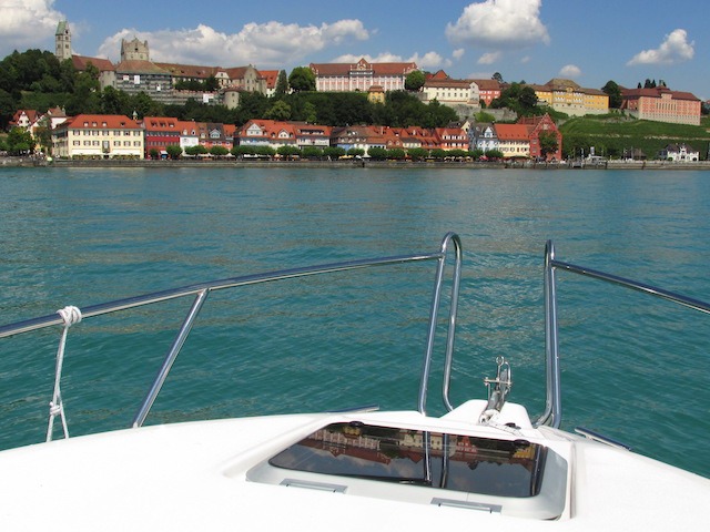 Das Bild zeigt im Hintergrund Meersburg am Bodensee, im Vordergrund sieht man den Bug des Bootes und das blaue Wasser des Bodensees. Die Stadt Meersburg spiegelt sich in der Luke des Boots.