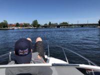 Das Foto wurde vom Fahrerstand aus gemacht, man sieht den Bug der 20 -Yachtline-. Hier liegt der Sohn der Familie und genießt die Sonne er trägt eine schwarz melierte kurze Hose, einen grauen Pullover und eine dunkelblaue Cape mit der Englandfahne drauf. Im Hinter Grund sieht man einen Hafen mit vielen Booten und eine Brücke.