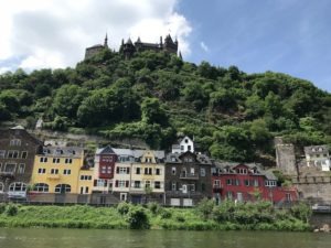 Auf dem Bild sieht man die Reichsburg von Cochem die Hoch oben auf dem Berg steht darunter befindet sich ein Wald und das Ufer des Rheins dort stehen schöne bunte Häuser.