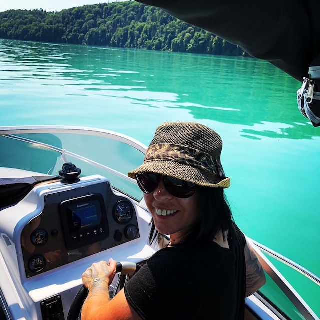 Frau H. steht am Steuer ihrer 20 -Yachtline-, sie trägt einen braunen Sommerhut und eine schwarze Sonnenbrille. Im Hintergrund sieht man türkis blaues Wasser und das bewaldete Ufer.