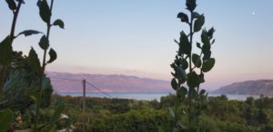 Das Bild wurde vom Festland aus gemacht, man sieht im Hintergrund die kroatische Berglandschaft und im Vordergrund grüne Büsche und Sträucher. Die Sonne geht gerade unter und der Himmel färbt sich in rosa, lila, blau, orange.