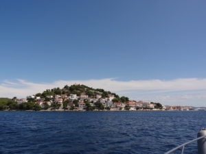 Blick vom Boot auf das Ufer Kroatiens. Blaues Wasser und am Horizont das Ufer mit einer kleinen Stadt die sich den Hügel hinauf schlängelt.