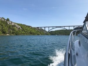 Seitlich aus der 23 -Yachtline- heraus fotografiert sieht man das Kroatischemeer, die steilen Küsten mit Baumen und Büschen und im Hintergrund sieht man eine große Brücke die zwei Inseln verbindet.