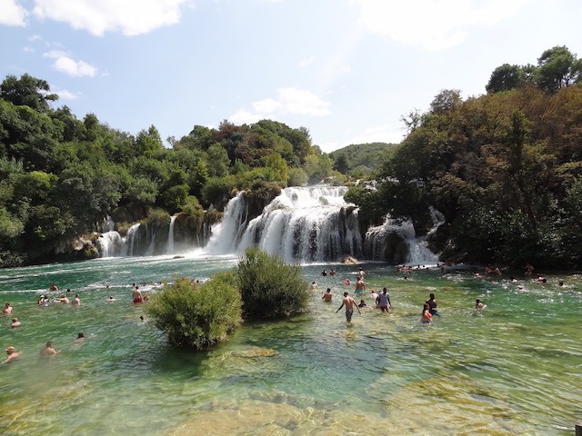 Das Foto zeigt die Krka Wasserfälle die zwischen Bäumen und Büschen liegen. Im Wasser sind viele Menschen die hier im türkis grünem Wasser baden.