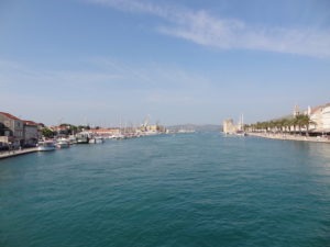 Links und recht die Stadt man sieht auf beiden Seiten viele Touristen die Kaimauern entlanglaufen. Links liegen einige kleiner Boote und Ausflugsschiffe im Hafen. Das Wasser in der Mitte ist türkisblau und es ist ein herrlicher Tag.