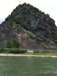Man sieht das steinige Ufer des Rheins, ein Schild mit der Aufschrift Loreley und dahinter den Imposanten Felsen.