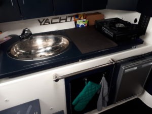 Man sieht den Küchenbereich der SR30 -Yachtline- die Familie hat auf der Arbeitsfläche neben dem Waschbecken einen tragbaren Kocher stehen, darunter befindet sich der silberne Einbaukühlschrank und ein großes Staufach.