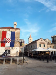 Man sieht die Innenstadt von Vodice, auf der rechten Seite ein kleines Restaurant und auf der linken Seite steht vor dem Haus eine Bühne hier stehen Instrumente am Haus hängt eine riesige kroatische Flagge.