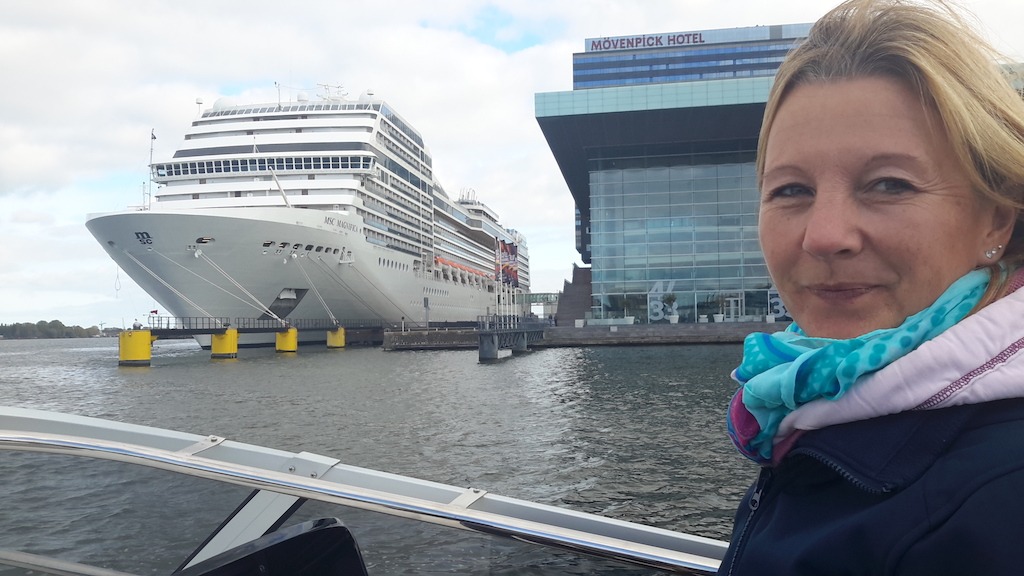 Das Bild zeit Frau T. wie sie am Steuerstand steht und in die Kamera lächelt, im Hintergrund sieht man das riesige Kreuzfahrtschiff MSC Magnifica und das Mövenpickhotel.
