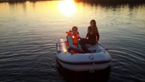 Das Foto wurde in der Dämmerung gemacht, es zeigt die zwei Töchter der Familie die im Schlauchboot sitzen. Die kleinere der beiden trägt eine neon orange Schwimmweste und ein Brille. Die Sonne spiegel sich hinter den Beiden im Wasser und lässt das Wasser gelb orange leuchten.