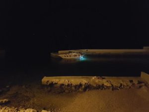 Das Foto wurde vom steinigen Strand aus gemacht, man sieht in der Ferne die SR30 -Yachtline an einer Kaimauer liegen. Es ist dunkle Nacht somit erkennt man nur durch das Licht der Laterne das Boot. Die Unterwasserbeleuchtung ist an und man kann erahnen das das Wasser türkis blau ist.