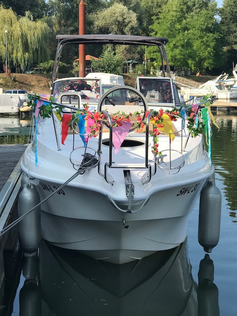 Die SR30 -Yachtline- der Familie liegt an einem Steg, links und recht hängen Fender an der Reling herunter. Das Boot wird an diesem Tag auf den Namen Happy getauft est ist mit vielen bunten Fahnen und Ästen mit Blüten geschmückt.